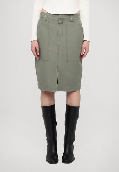 Falda de algodón color caqui con una abertura delantera, dos grandes bolsillos, presillas para cinturón y una silueta recta, combinada con botas de cuero negras hasta la rodilla.
