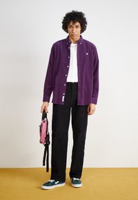 Paarse langemouwen shirt, witte t-shirt eronder, zwarte broek, groene schoenen, een kleine roze rugzak vasthoudend. Glad materiaal, met knopen ontwerp.