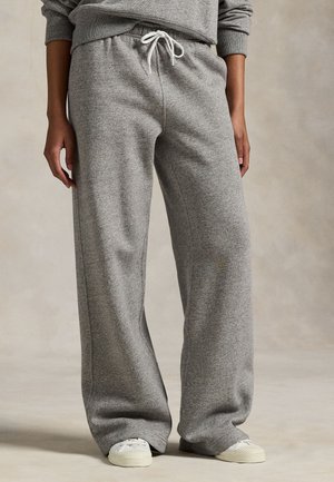 Person som har på seg løstsittende grå joggebukser med snøring i midjen og hvite sneakers, står mot en nøytral bakgrunn.