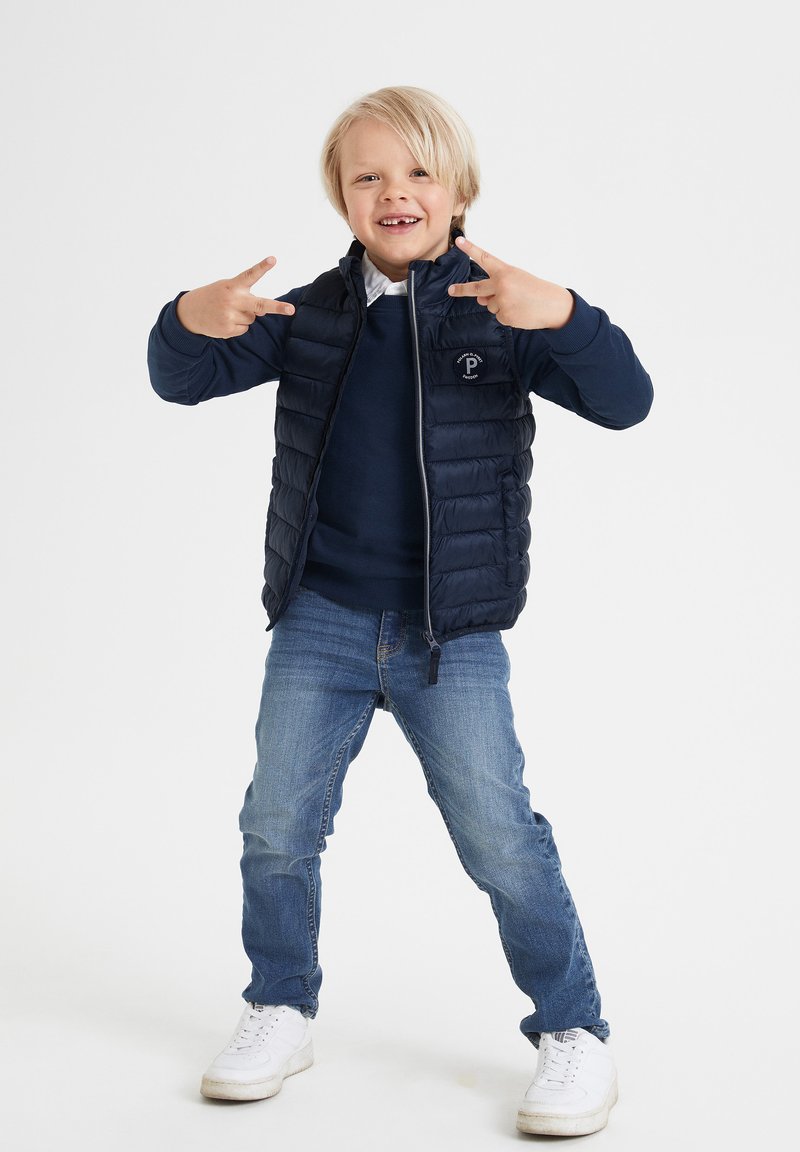 Marineblaue Puffer-Weste mit horizontalem Steppmuster über einem dunklen Sweatshirt, kombiniert mit hellblauen Jeans und weißen Sneakern.