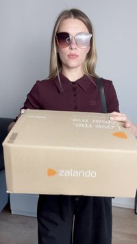 Caixa de cartão da Zalando com o logótipo laranja, segura por uma pessoa que usa óculos de sol oversized, uma camisa de colarinho em bordeaux escuro e calças pretas.