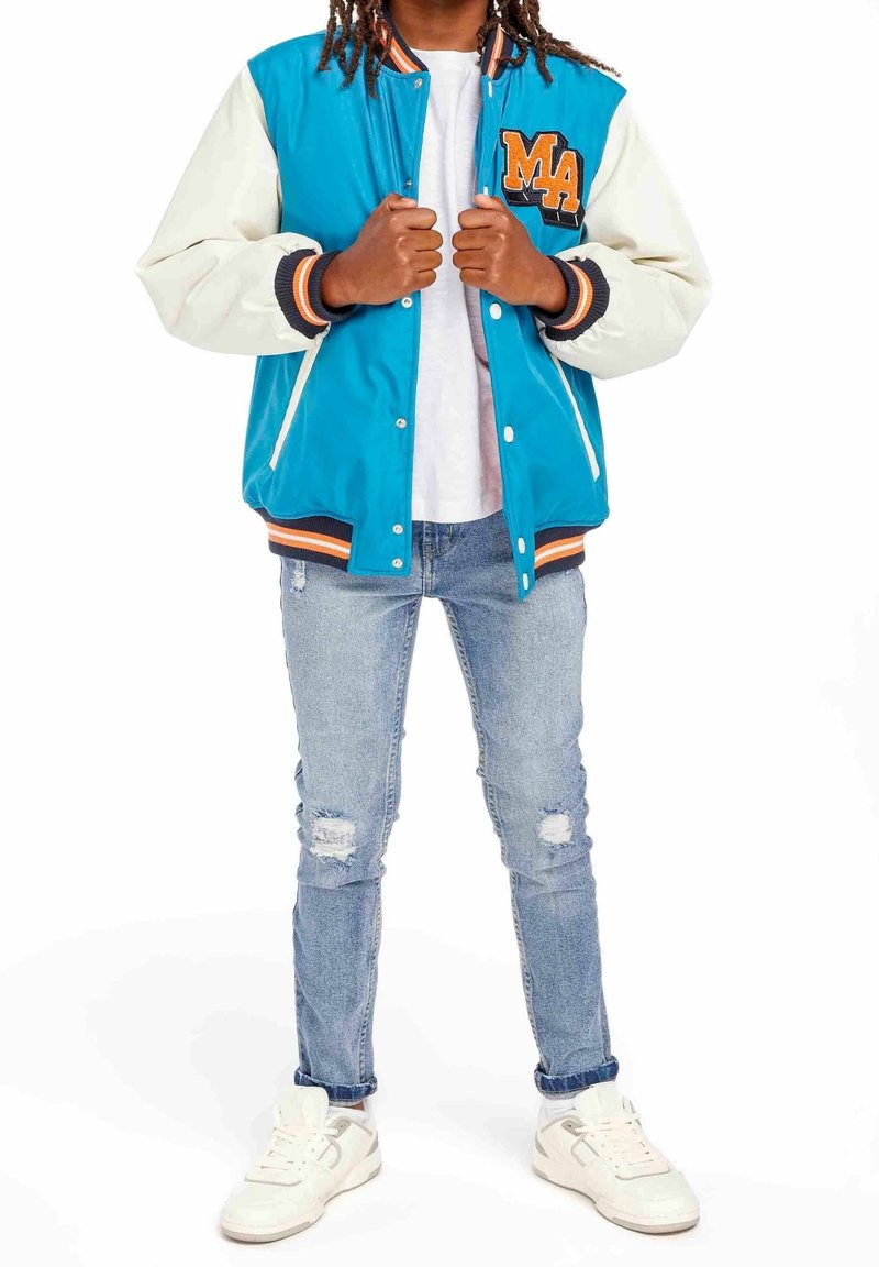 Jeune personne portant une veste universitaire bleue et blanche, t-shirt blanc, jean skinny déchiré et baskets blanches, debout les mains tenant le devant de la veste.