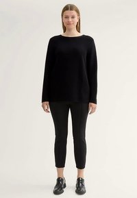 Svart stickad tröja med långa ärmar, rund halsringning och lös passform, kombinerad med svarta smala byxor och svarta snörskor.