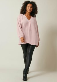 Rosa Langarmbluse mit V-Ausschnitt und Falten, kombiniert mit schwarzen Lederleggings und Stiefeletten. Weicher, strukturierten Stoff.