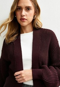 Burgundy knit cardigan with ribbed texture and wide sleeves, worn over a white top. Features a relaxed fit and open front design.