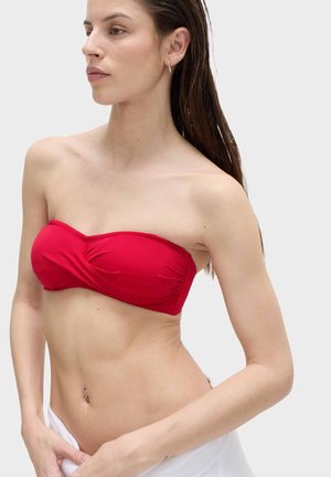 Kobieta z mokrymi włosami, ubrana w czerwony bikini bez ramiączek i białe majtki, pozująca z ręką przy talii na jednolitym tle.