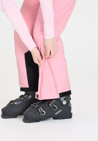 Rosa skidbyxor med dragkedja och kardborreband vid ankeln, i kombination med svarta skidstövlar med justerbara spännen och texturerade mönster.