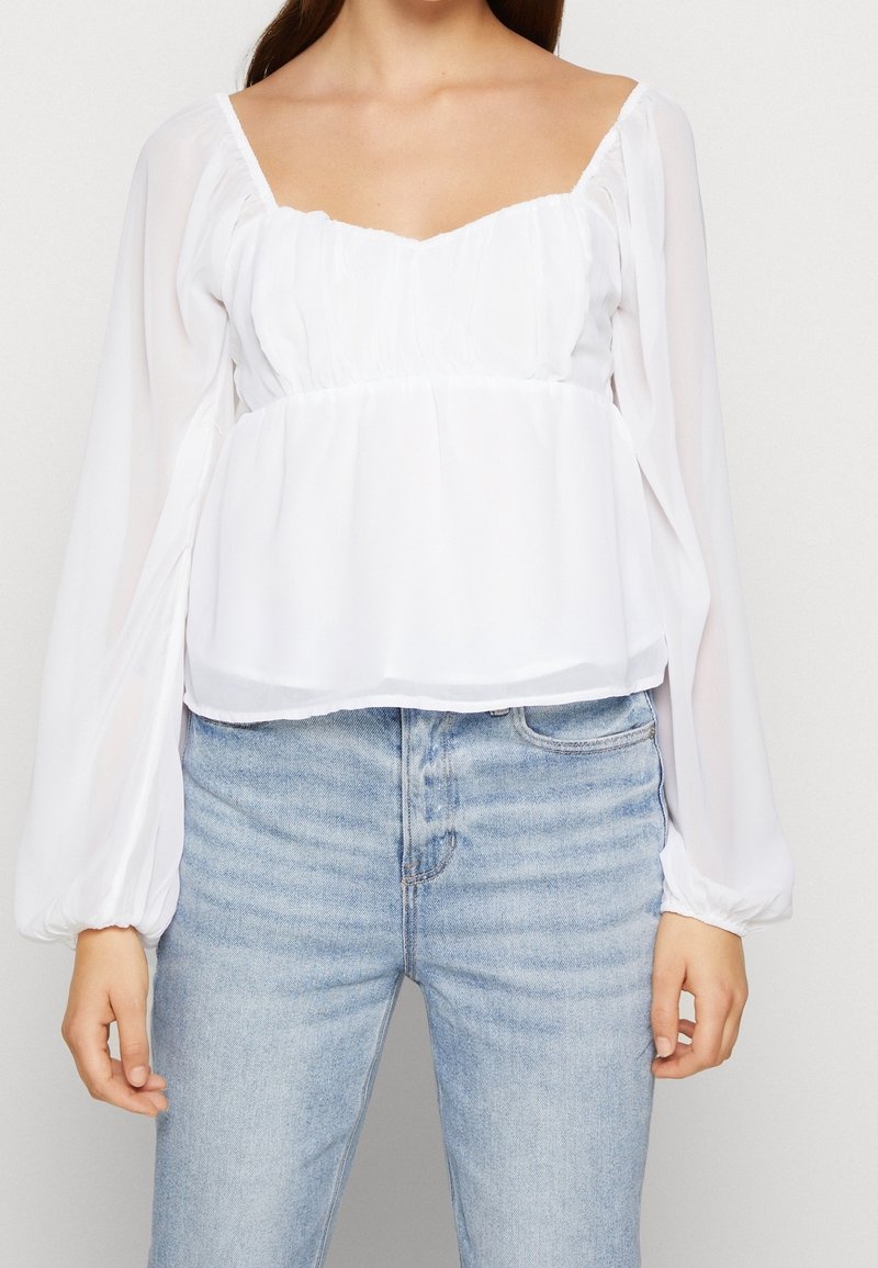 Femme portant un chemisier blanc transparent à manches longues avec un bustier froncé et un jean taille haute bleu clair sur fond uni.