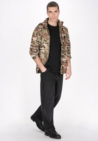 Kamouflagejacka i nyanser av grönt och brunt, svart T-shirt, svarta jeans och robusta svarta stövlar; avslappnad passform med texturerad tyg.
