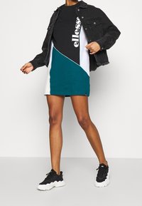 Veste en denim noire portée sur une robe bicolore noire et bleu sarcelle avec un ourlet arrondi. Accompagnée de baskets noires à semelle blanche.