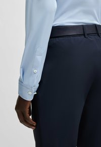 Chemise bleu clair à manches longues avec des poignets à boutons, associée à un pantalon marine sombre. La chemise présente une texture subtile et une coupe ajustée.