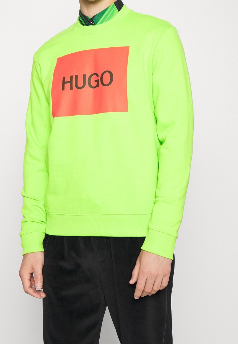 Felpa verde neon con scollo a girocollo, caratterizzata da un audace design rettangolare rosso con testo nero "HUGO" al centro. Tessuto morbido.