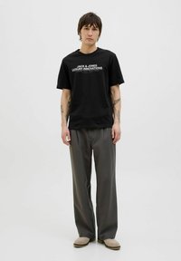 Svart kortärmad t-shirt med vit text "JACK & JONES LUXURY INNOVATIONS," i kombination med grå byxor och beige slip-on skor.