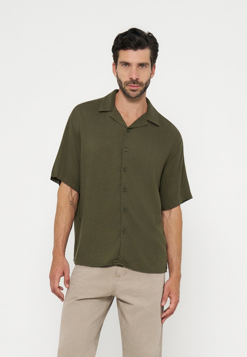 Homme aux cheveux foncés portant une chemise boutonnée à manches courtes vert olive et un pantalon beige, posant devant un fond uni.