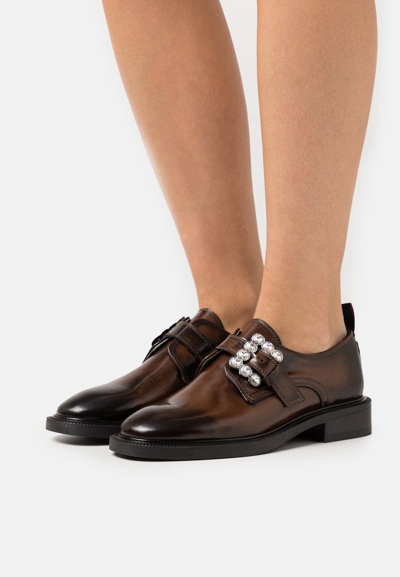 Chaussures à boucles moniales en cuir marron avec de grandes boucles décoratives argentées, portées pieds nus sur un fond blanc.