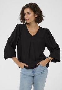Blouse noire à col en V avec des manches bouffantes, en tissu texturé et à coupe décontractée. Associée à un jean bleu clair, elle présente un design simple et moderne.