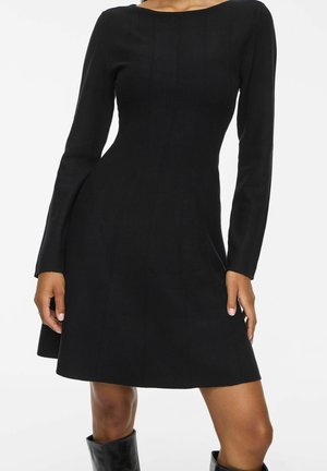 Robe noire moulante à manches longues avec des coutures verticales subtiles, portée avec des bottes noires mi-mollet, montrée des épaules aux genoux.
