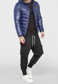 Mörkblå vadderad jacka med quiltad textur och huva, kombinerad med svarta cargobyxor med sidofickor. Vita sneakers fulländar outfiten.