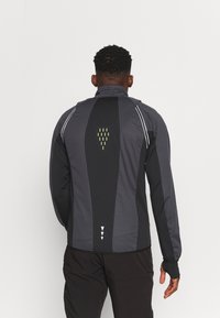 Veste de sport noire et grise avec un col montant, des accents réfléchissants et un dos à motifs avec des détails vert clair. Manches longues avec trous pour les pouces.