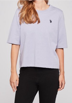 Femme portant un t-shirt à manches courtes violet clair avec un petit logo de cheval noir sur le côté gauche de la poitrine et un pantalon noir.