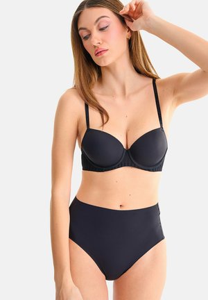 Femme ajustant ses cheveux, portant un soutien-gorge noir rembourré assorti et une culotte taille haute sur un fond uni.