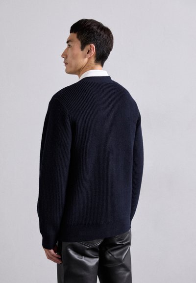 Tumes sinine sooniknitted sviiter ümmarguse kaeluse ja pikad varrukatega, koos mustade nahkpükstega, nähtud tagant.