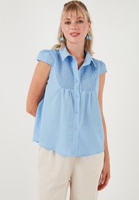 Blusa azzurra chiara a maniche corte con chiusura a bottoni frontale, dettagli in pizzo sullo scollo e una vestibilità rilassata con orlo curvo.