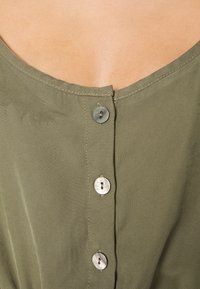 Blouse en tissu vert olive avec un col arrondi et cinq boutons ronds de couleur claire sur le devant, avec des détails de coutures visibles.