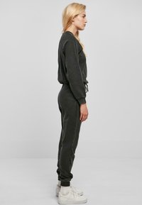 Mørk grå, langermet jumpsuit med tilpasset midje, rillede mansjetter og snøring. Kombinert med hvite sneakers og synlige ankelsokker.
