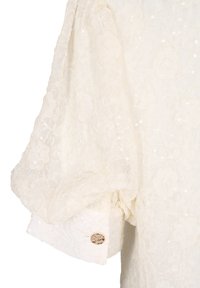 Blouse en dentelle blanche avec des motifs floraux, tissu texturé, manches oversized et un accent de bouton doré sur le poignet.