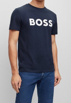 Uomo che indossa una maglietta blu navy con la scritta bianca "BOSS" e jeans blu, in piedi davanti a uno sfondo grigio chiaro uniforme.