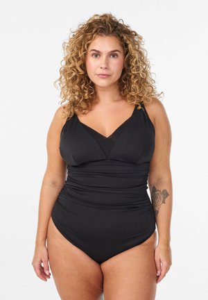 Proua lokkidega naine, kes kannab musta ühes tükis ujumisriietust, näitab vasakul küünarvarrel roositätoveeringut ja vaatab otse ette.