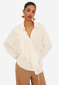Blusa color crema con dettagli arricciati sul davanti, colletto con bottoni e vita con cordoncino. Tessuto leggero e vestibilità comoda.