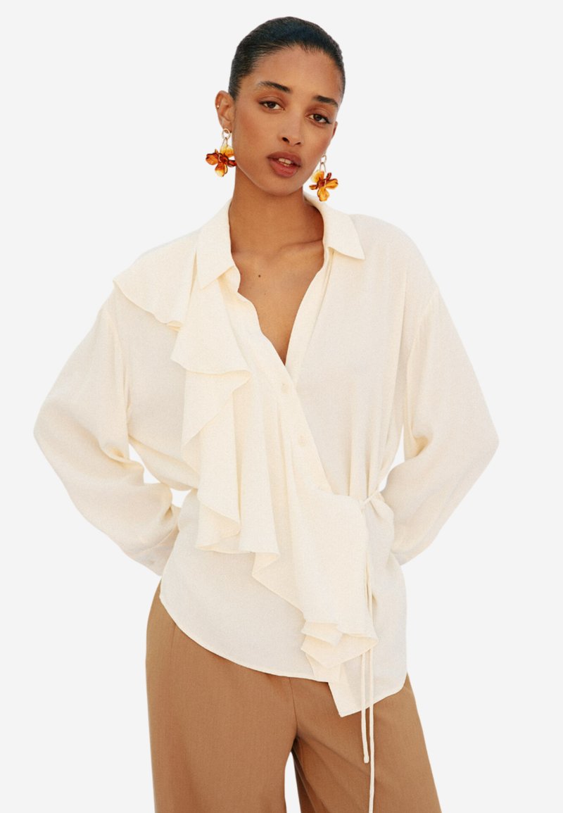 Blusa color crema con dettagli arricciati sul davanti, colletto con bottoni e vita con cordoncino. Tessuto leggero e vestibilità comoda.