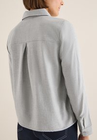 Szara koszula zapinana na guziki z kołnierzykiem, wykonana z teksturowanej tkaniny, z centralnym plisowaniem z tyłu oraz długimi rękawami zakończonymi mankietami zapinanymi na guziki.