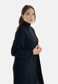 Cardigan nero lavorato a maglia con un motivo testurizzato, apertura frontale e maniche lunghe. Presenta un collo alto a coste sotto per stratificare.