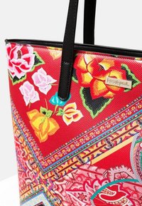 Sac fourre-tout rouge avec motifs floraux, finition texturée et poignées noires. Présente un logo métallique et des détails géométriques vibrants.