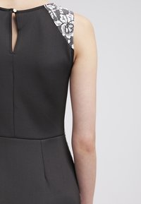 Zwarte jurk met een gestructureerde zwart-wit patroon op de schouder. Heeft een keyhole rugontwerp en gestikte naden. Gladde stofafwerking.