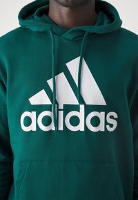 Sudadera verde de algodón con un gran logo blanco de Adidas. Incluye un bolsillo tipo canguro y capucha ajustable con cordón.