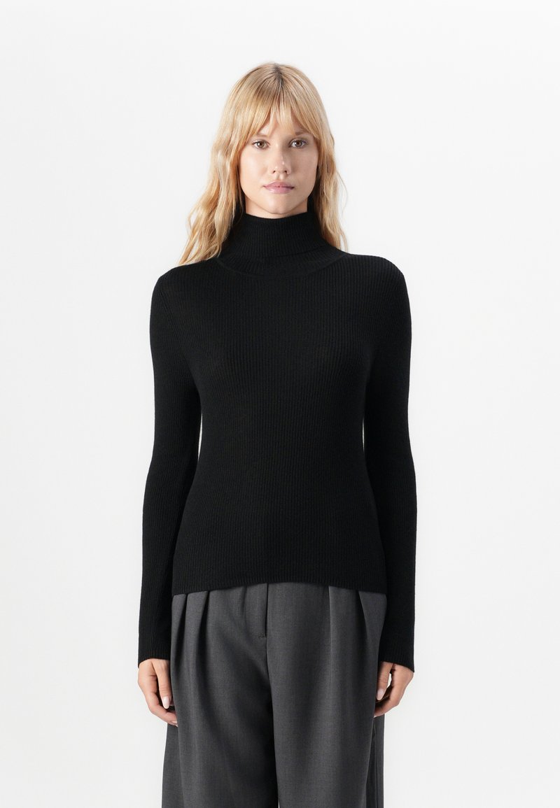 Pull à col roulé noir à côtes avec manches longues, fabriqué en tissu texturé, associé à un pantalon gris plissé.
