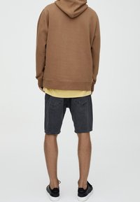 Sudadera marrón con capucha, camiseta amarilla debajo, pantalones cortos de mezclilla gris y zapatillas negras. Corte casual y relajado con costuras visibles.