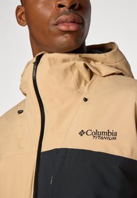 Beige og svart Columbia Titanium jakke med glidelås foran, justerbar hette og logodetalj på brystet. Glatt stofftekstur.