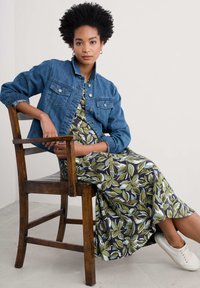 Veste en denim avec des boutons argentés, portée sur une robe maxi fleurie avec des feuilles vertes sur un fond sombre. Des baskets décontractées complètent le look.