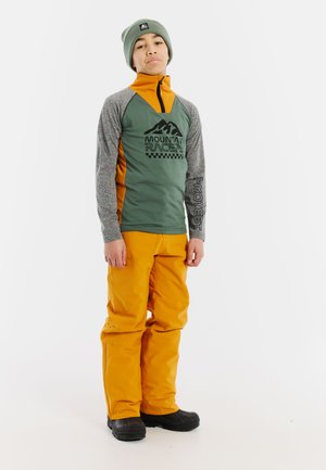 Haut thermique superposé vert et orange avec un logo, manches grises et pantalon de neige orange. Des bottes d'hiver noires complètent la tenue.
