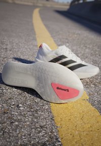 Бели маратонки Adidas с черни ивици, лежащи на асфалтов път с жълта линия, показващи подметка с червен надпис Continental.