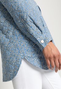 Blauw bloemenpatroon shirt met pofmouwen, een knoopsluiting bij de manchetten en een afgeronde onderkant, gecombineerd met witte broek.