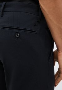 Κοντινό πλάνο σκούρων υφασμάτινων παντελονιών με τσέπη στο πίσω μέρος που κλείνει με κουμπί και μικρό λογότυπο κοντά στην τσέπη, φορεμένα από άτομο με ορατό χέρι.