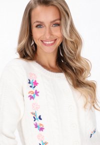 Biały kardigan z dzianiny w warkocze z kolorowym haftem kwiatowym po lewej stronie, z okrągłymi guzikami i miękką fakturą.