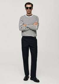 Gestreiftes Langarmhemd mit navy-blauen und weißen Streifen, kombiniert mit schwarzen Hosen und schwarzen Schuhen. Modell trägt dunkle Sonnenbrille und hat die Arme verschränkt.