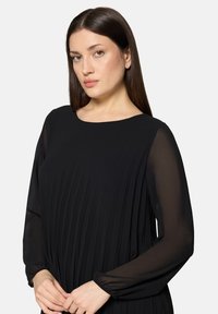 Czarna plisowana sukienka z długimi, przejrzystymi rękawami, okrągłym dekoltem i subtelną fakturą, prezentująca płynącą sylwetkę i gładką tkaninę.